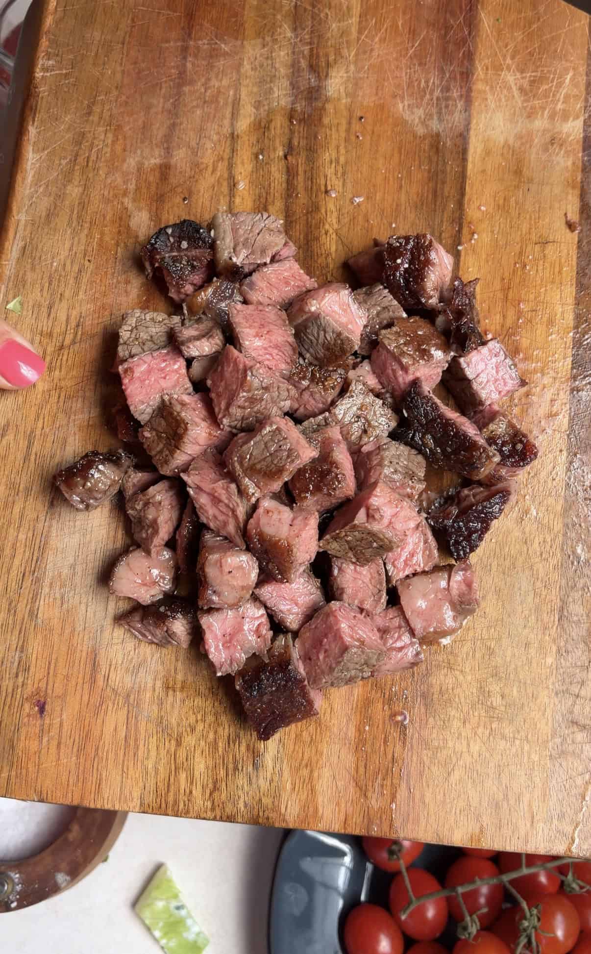 Cubed pieces of cooked steak rest on a wooden cutting board. A hand with pink-painted nails is visible to the left, and cherry tomatoes sit on a blue plate at the bottom right corner.