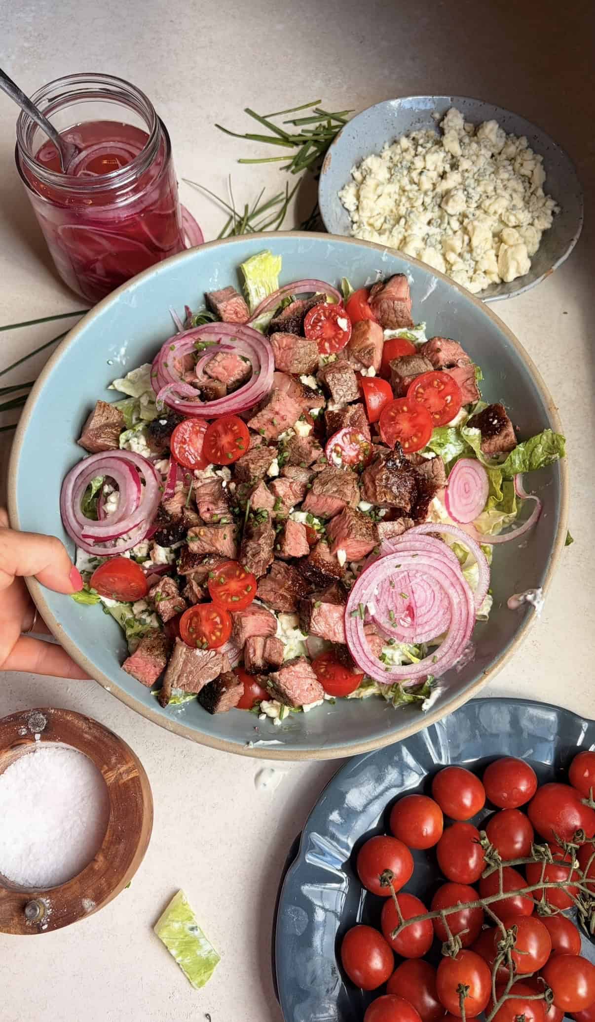 A hand holds a bowl of steak salad with cherry tomatoes, red onions, and lettuce. Nearby are a jar of pickled onions, a bowl of crumbled cheese, cherry tomatoes, and a wooden salt container.