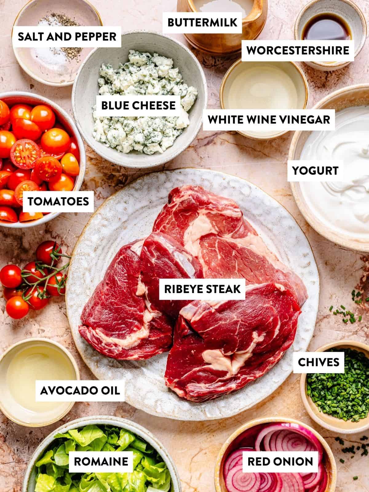Two raw ribeye steaks on a plate, surrounded by bowls of ingredients—cherry tomatoes, blue cheese, chives, red onion, romaine, avocado oil, yogurt, buttermilk, Worcestershire sauce, white wine vinegar, salt, and pepper—perfect for preparing a delicious blue cheese and steak salad.