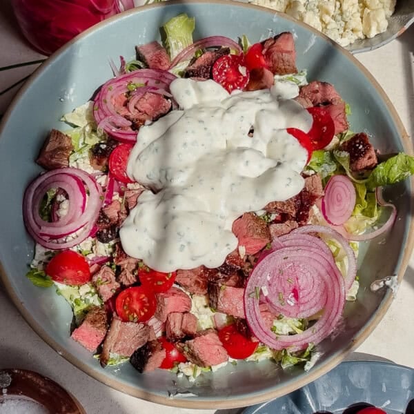 A bowl of blue cheese and steak salad with lettuce, cherry tomatoes, sliced red onions, steak pieces, crumbled cheese, and a large dollop of creamy blue cheese dressing on top.