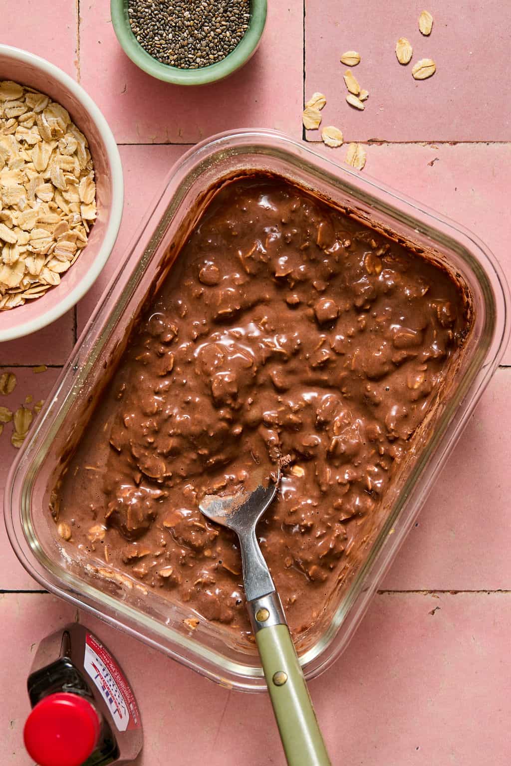 A glass container filled with brownie overnight oats and a spoon, surrounded by a bowl of oats, chia seeds, and a bottle of vanilla extract on a pink tiled surface.