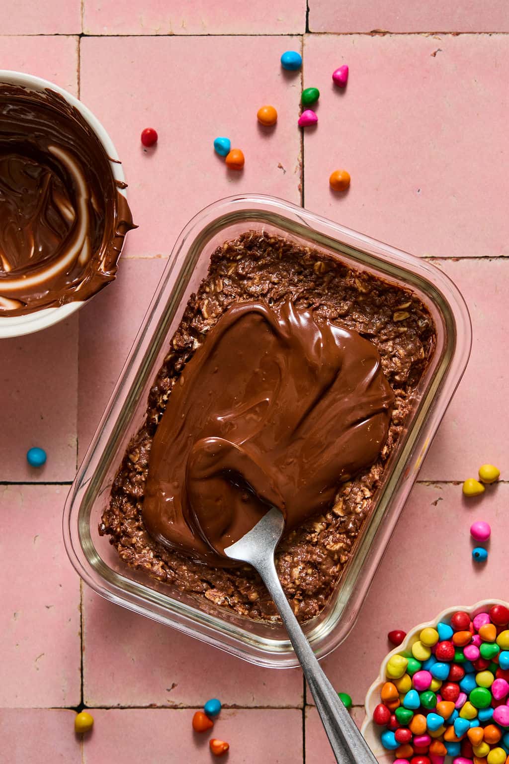 A glass dish of brownie overnight oats and chocolate oat mixture is being spread with melted chocolate by a spoon. Colorful candy sprinkles and a bowl with more melted chocolate are nearby on a pink tiled surface.