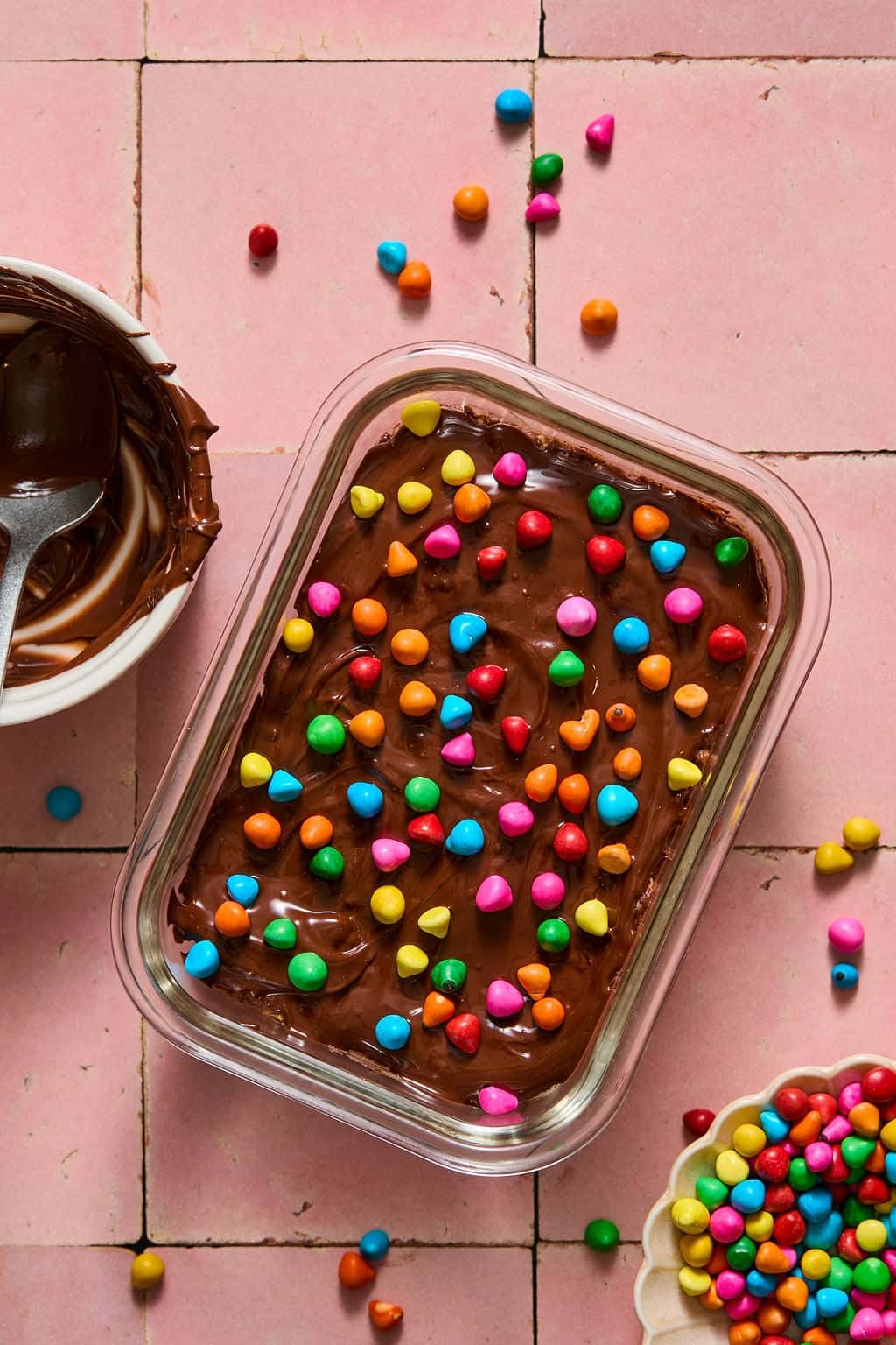 A glass container holds brownie overnight oats, topped with colorful candy-coated chocolates. Nearby are a bowl with melted chocolate and a bowl of candy pieces on a pink tiled surface. Some candies are scattered around.