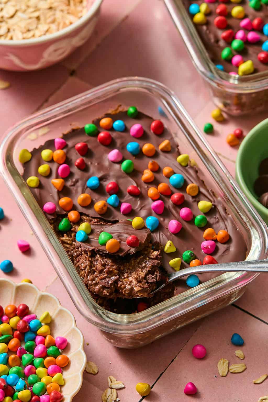 A glass dish filled with brownie overnight oats is topped with colorful round candy sprinkles. A spoon rests inside, with some dessert scooped. Scattered sprinkles and oats are visible on the pink surface around the dish.