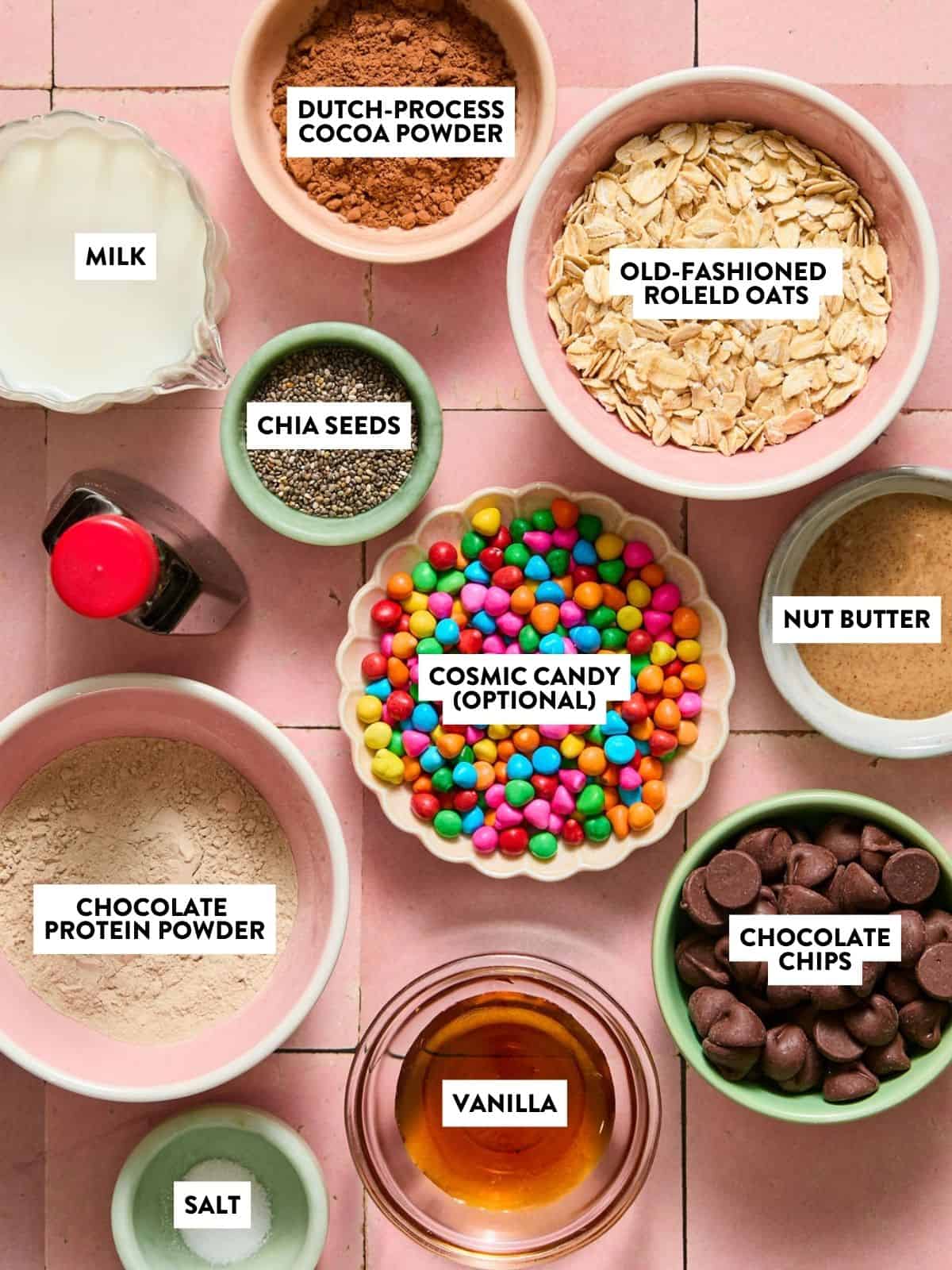 A top view of overnight oats ingredients in bowls on a pink tiled surface, including oats, nut butter, vanilla, chocolate chips, milk, cocoa powder, chocolate protein powder, salt, chia seeds, and colorful candy.