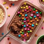 A glass dish of rich chocolate brownie overnight oats topped with colorful candy pieces and partially scooped out with a spoon. Surrounding bowls contain oats and other ingredients, all set on a pink tiled surface.