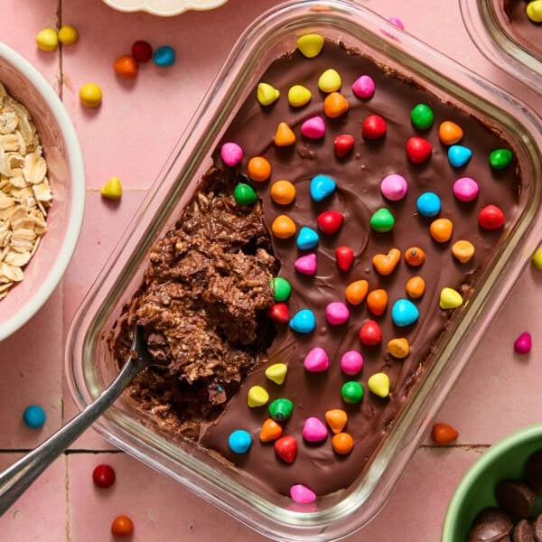 A glass dish of rich chocolate brownie overnight oats topped with colorful candy pieces and partially scooped out with a spoon. Surrounding bowls contain oats and other ingredients, all set on a pink tiled surface.