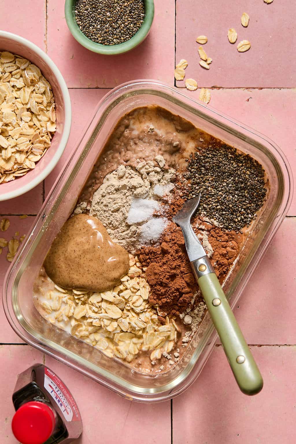A glass container with brownie overnight oats made from oats, cocoa powder, chia seeds, almond butter, and protein powder, with a green-handled spoon. Nearby are bowls of oats, chia seeds, and a bottle of vanilla. All on a pink tiled surface.