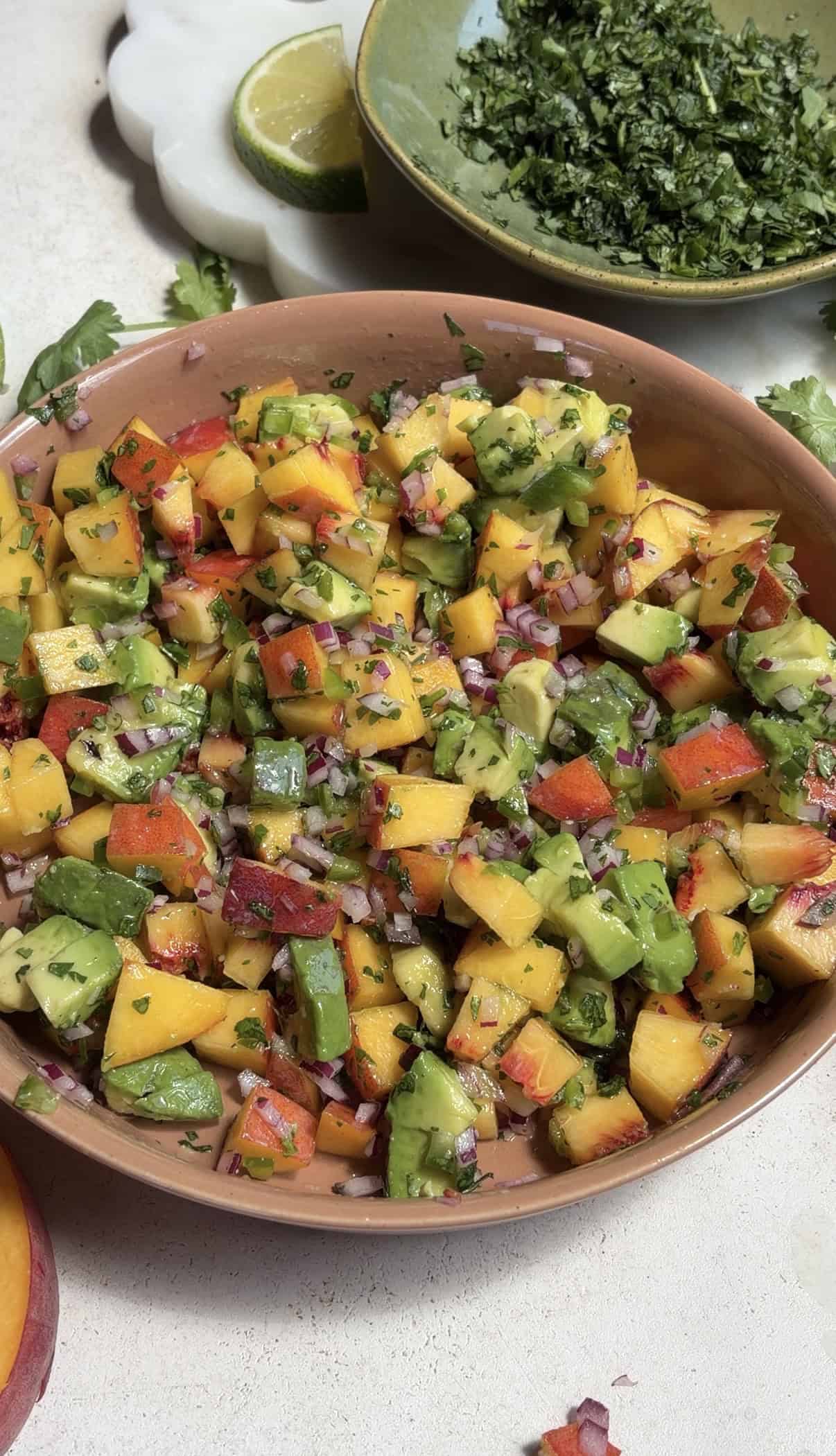 A bowl of colorful peach salsa with chopped avocado, red onion, cilantro, and red bell pepper sits on a light surface. A green plate with chopped herbs and a lime are in the background.