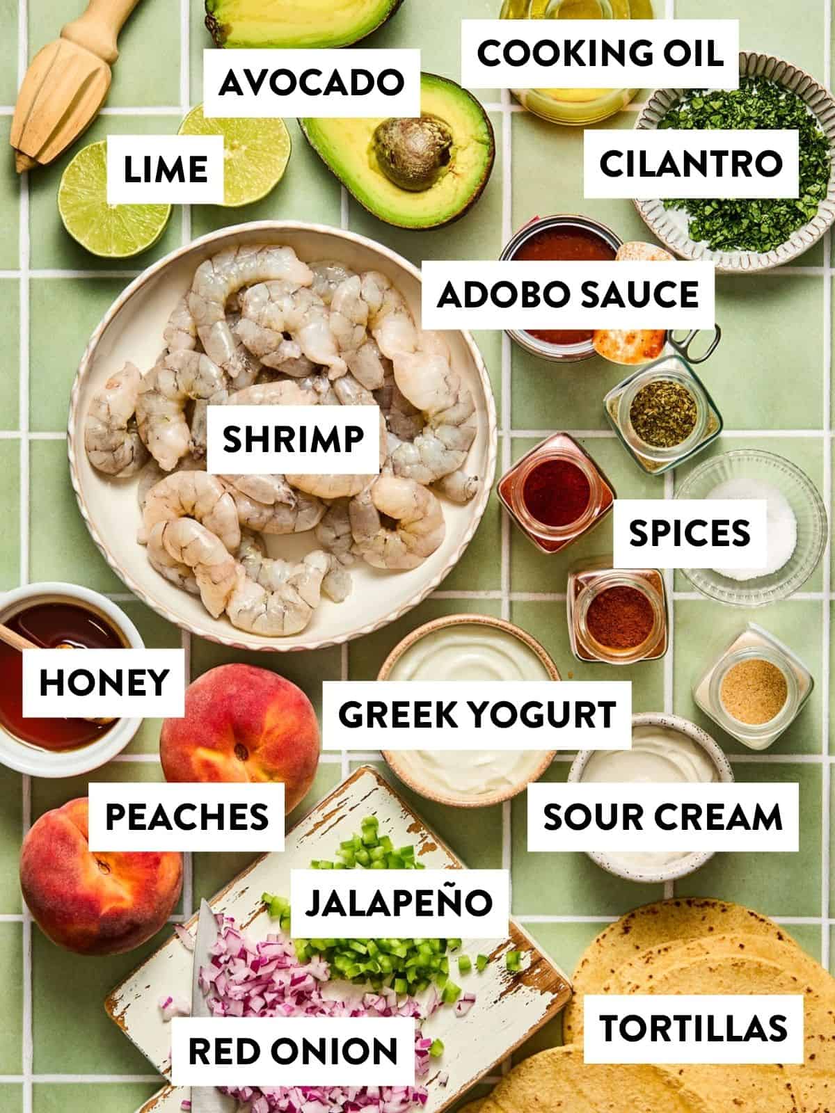 Various ingredients labeled, including shrimp, avocado, lime, cooking oil, cilantro, adobo sauce, spices, honey, peaches, Greek yogurt, sour cream, jalapeño, red onion, and tortillas on a tiled surface—everything you need to prepare vibrant chipotle shrimp tacos.