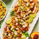 Five chipotle shrimp tacos topped with peach salsa, creamy sauce, and cilantro are arranged on a white platter with lime wedges. A side bowl of salsa and a peach slice are visible next to the tacos.