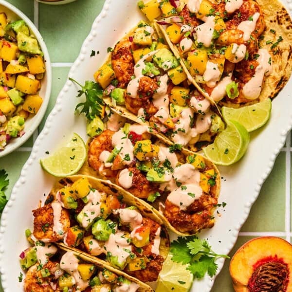 Five chipotle shrimp tacos topped with peach salsa, creamy sauce, and cilantro are arranged on a white platter with lime wedges. A side bowl of salsa and a peach slice are visible next to the tacos.