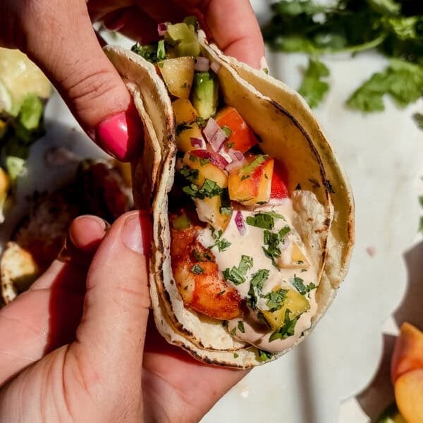 A person holding a chipotle shrimp taco filled with peach salsa and creamy sauce in a corn tortilla.