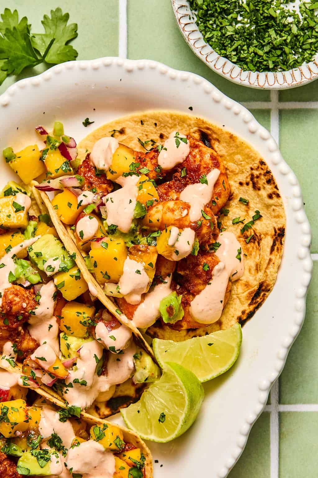 Two chipotle shrimp tacos on a plate are topped with diced mango, avocado, salsa, creamy sauce, and fresh herbs, served with lime wedges. A small bowl of chopped cilantro is visible in the background.