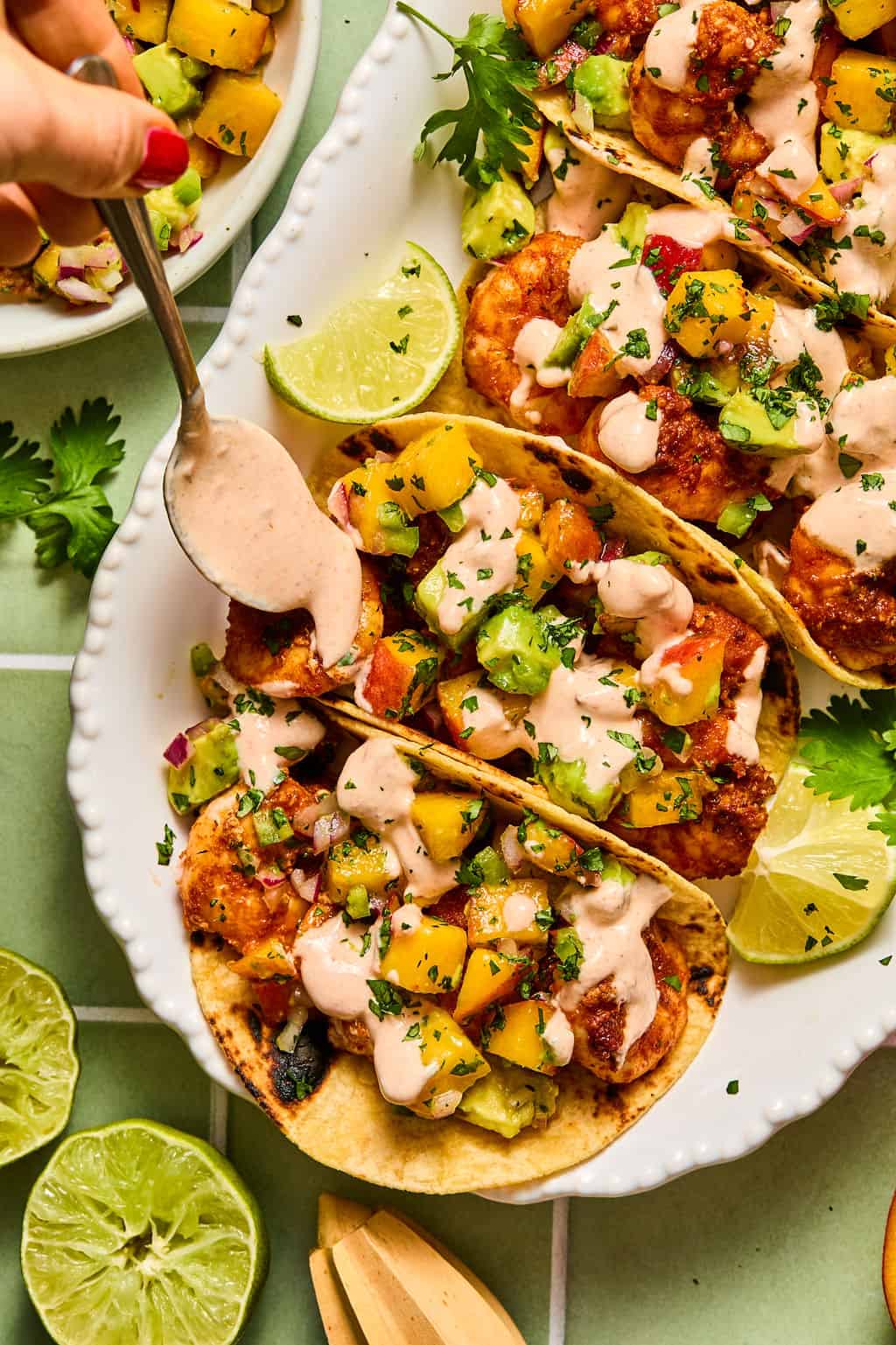 Three tacos filled with crispy breaded shrimp, mango salsa, and creamy sauce are served on a white plate with fresh cilantro and lime wedges. A hand is drizzling sauce over the tacos with a spoon.