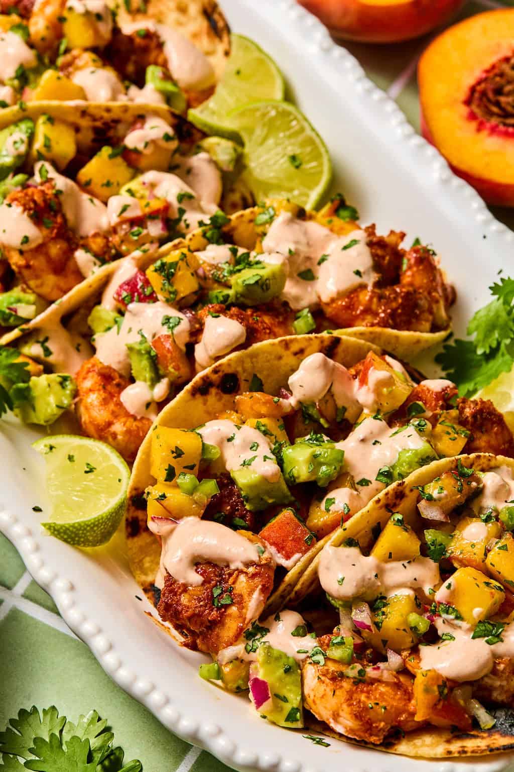 Three tacos filled with grilled shrimp, fresh salsa with avocado and peaches, and drizzled with creamy sauce, are served on a white platter with lime wedges and cilantro garnish.
