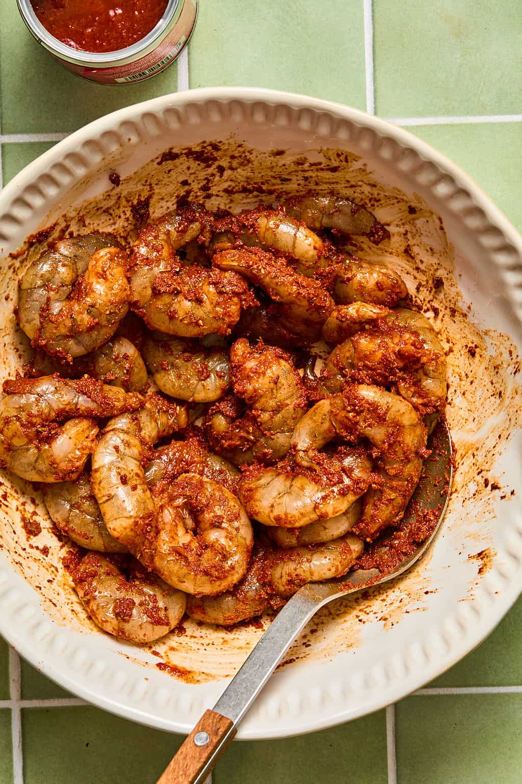 A white bowl of raw shrimp coated in a reddish marinade—perfect for chipotle shrimp tacos—sits on a green tiled surface with a spoon and a jar of sauce nearby.