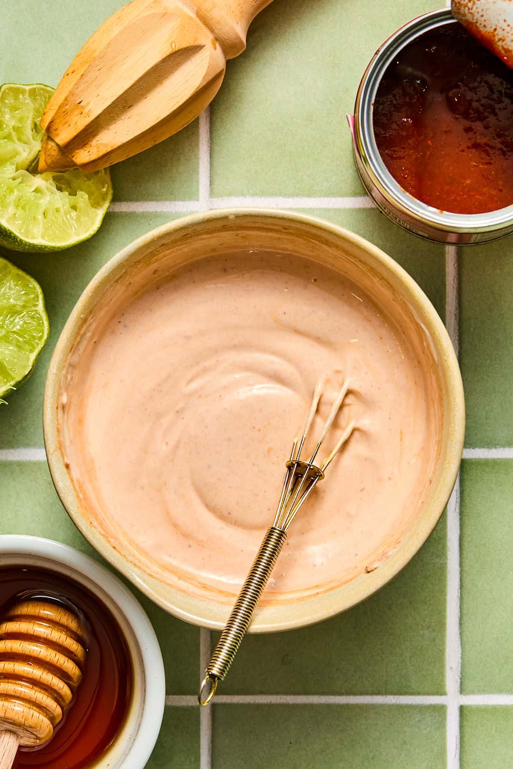 A bowl of creamy sauce with a small whisk, surrounded by a honey dipper in honey, cut limes, a wooden citrus reamer, and an open can of red sauce—perfect ingredients for making chipotle shrimp tacos—all arranged on a green tiled surface.