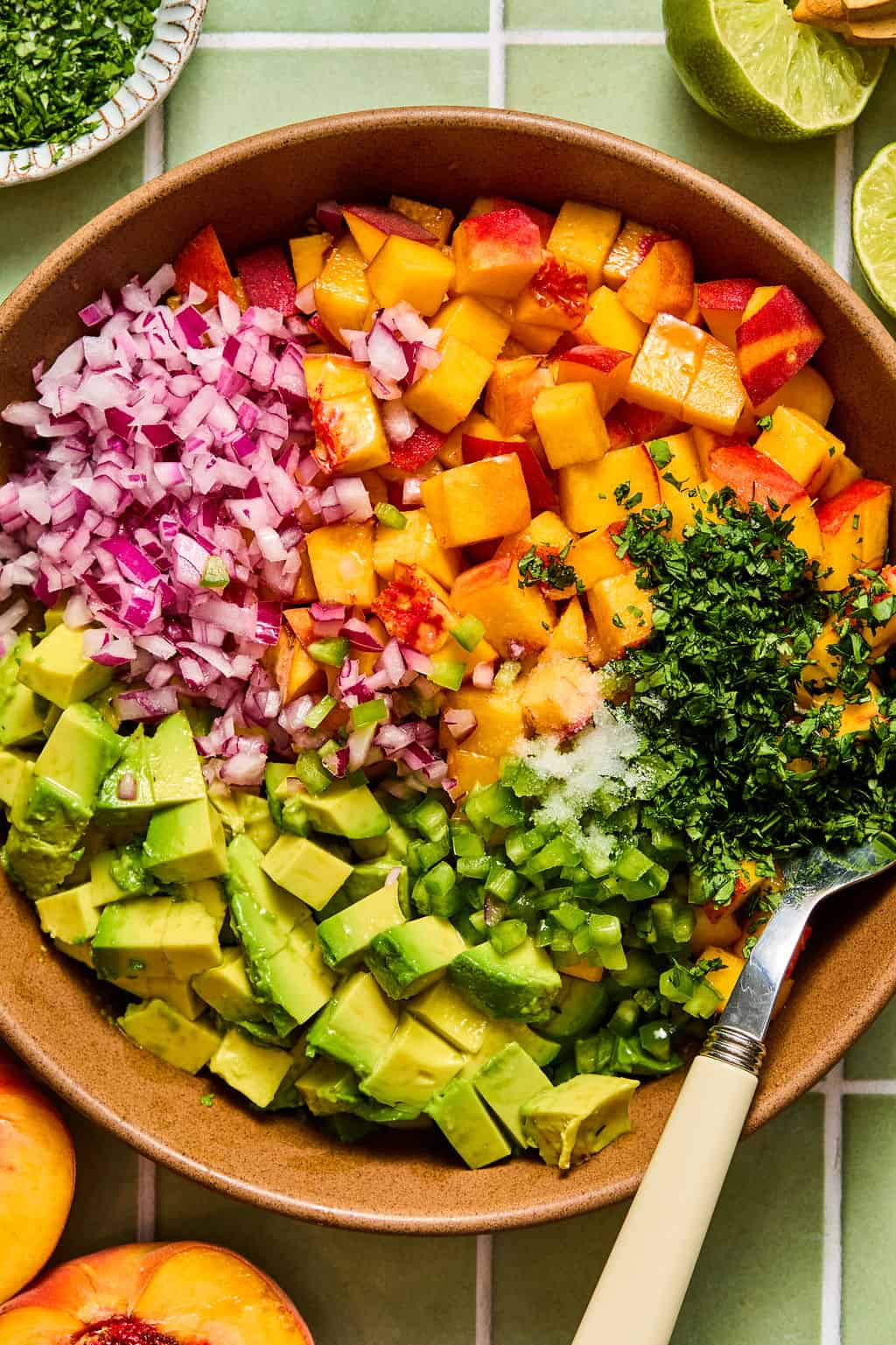 A bowl with diced avocado, peaches, red onion, cilantro, green pepper, and salt on a green tiled surface—perfect as a fresh topping for chipotle shrimp tacos—with a spoon and lime halves nearby.