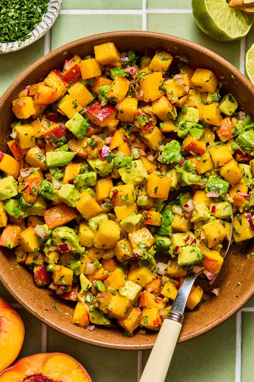 A bowl of fresh peach salsa with diced peaches, avocado, red onion, cilantro, and bell pepper, placed on a green tiled surface with lime wedges and herbs nearby—perfect for serving alongside chipotle shrimp tacos.