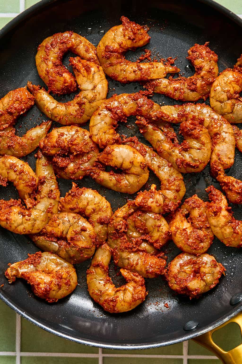 Raw shrimp coated in a red, spicy seasoning—perfect for chipotle shrimp tacos—are arranged in a single layer in a black skillet, ready to be cooked. The skillet sits on a green and white tiled surface.