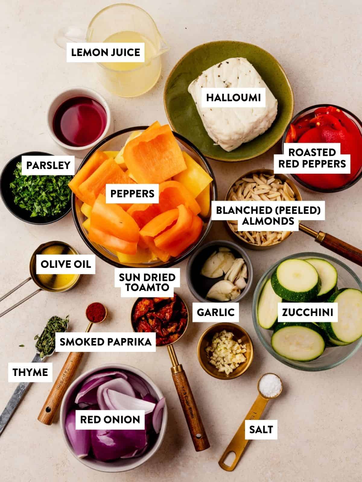 Various ingredients in bowls and plates, including halloumi, chopped peppers, zucchini, red onion, herbs, garlic, almonds, sun-dried tomato, roasted red peppers, olive oil, lemon juice, smoked paprika, thyme, and salt—perfect for assembling flavorful halloumi kebabs.