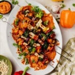 Grilled vegetable and halloumi kebabs with zucchini, bell peppers, and onions served over a bed of romesco sauce on a white platter, garnished with fresh herbs. Surrounding the platter are garlic, bell peppers, and a bowl of slivered almonds.