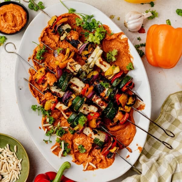 Grilled vegetable and halloumi kebabs with zucchini, bell peppers, and onions served over a bed of romesco sauce on a white platter, garnished with fresh herbs. Surrounding the platter are garlic, bell peppers, and a bowl of slivered almonds.