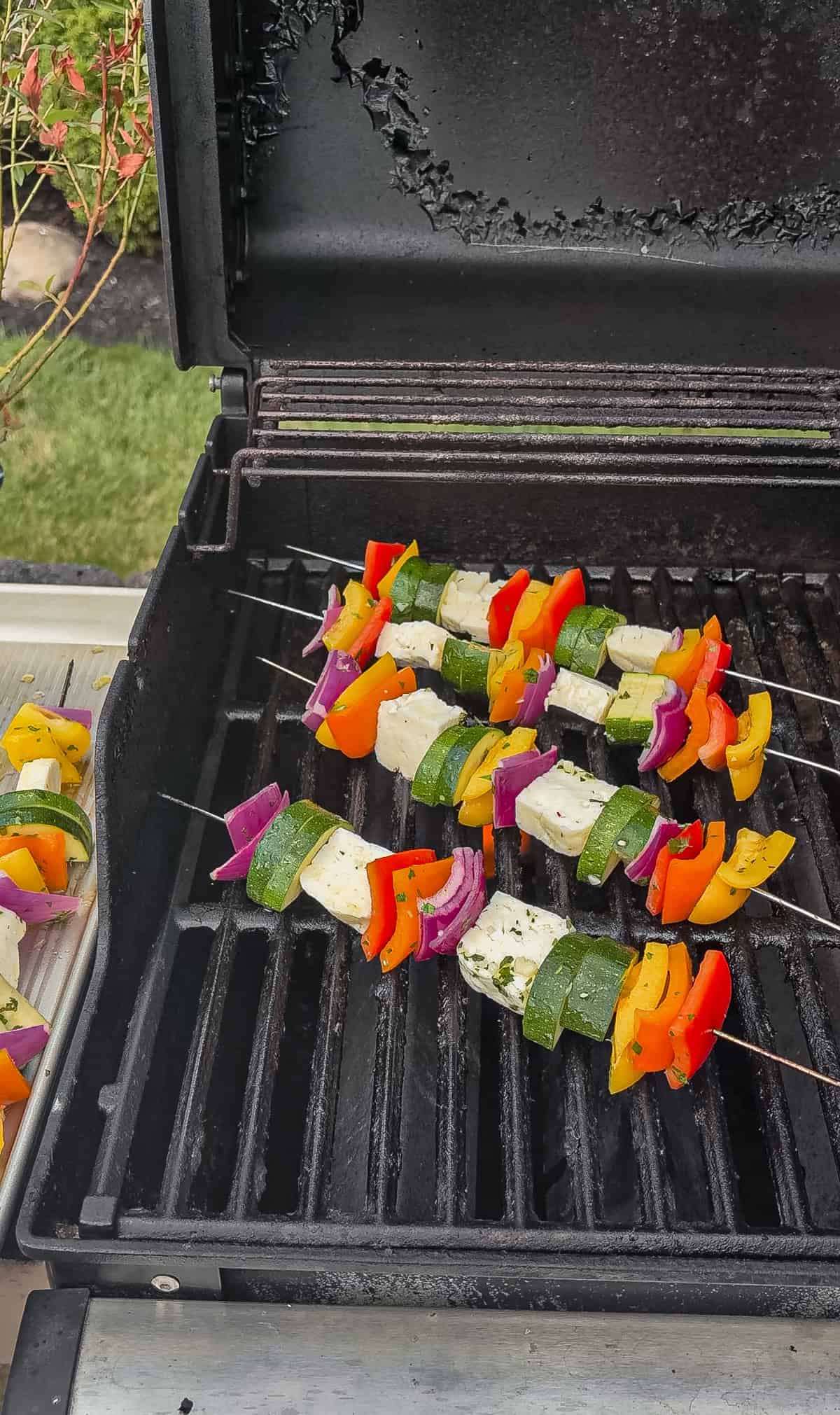 Colorful vegetable and halloumi skewers, including zucchini, bell peppers, red onion, and cubes of halloumi are grilling on an outdoor barbecue grill.