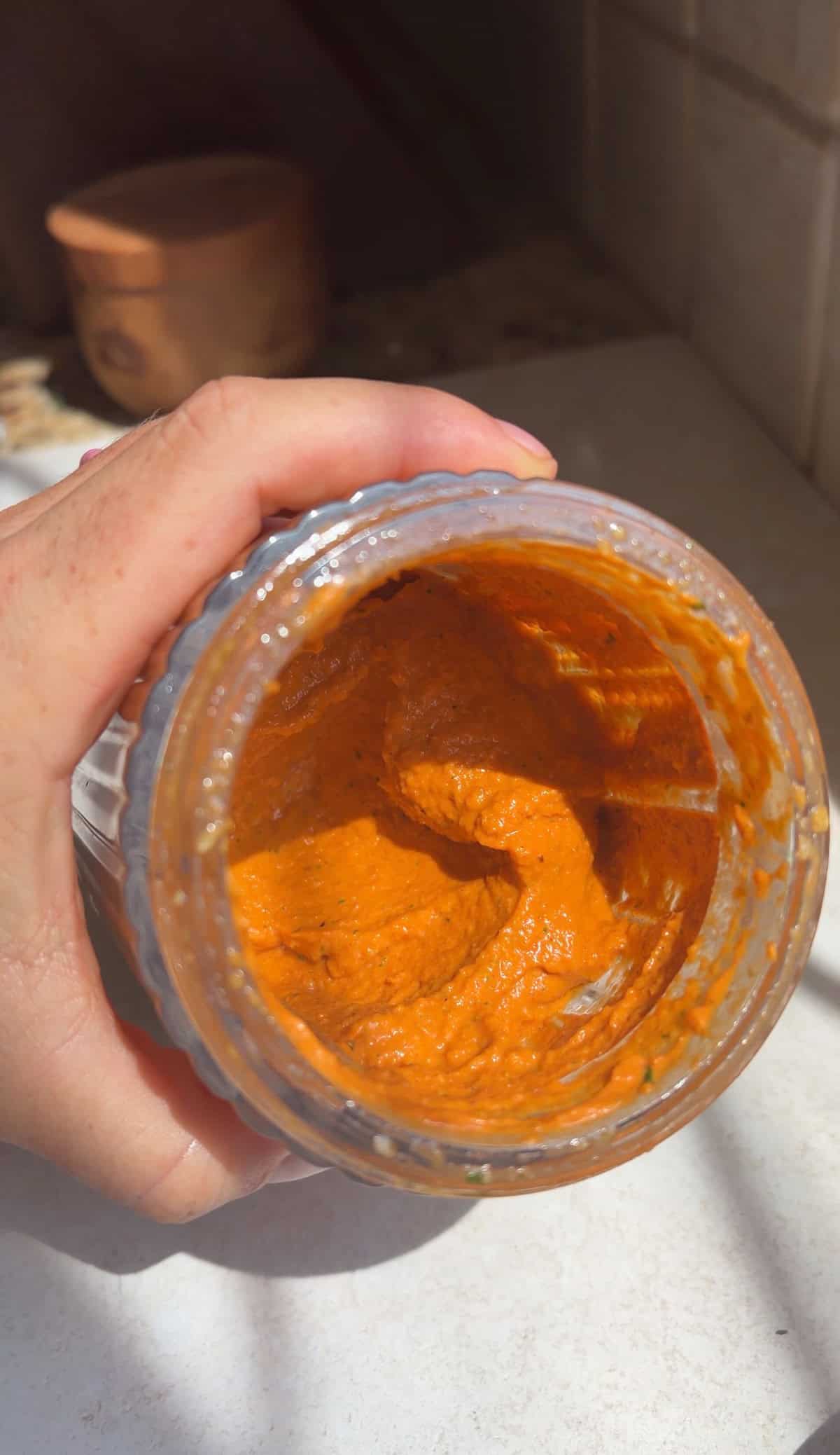 A hand holds an open glass jar containing a thick homemade romesco sauce.