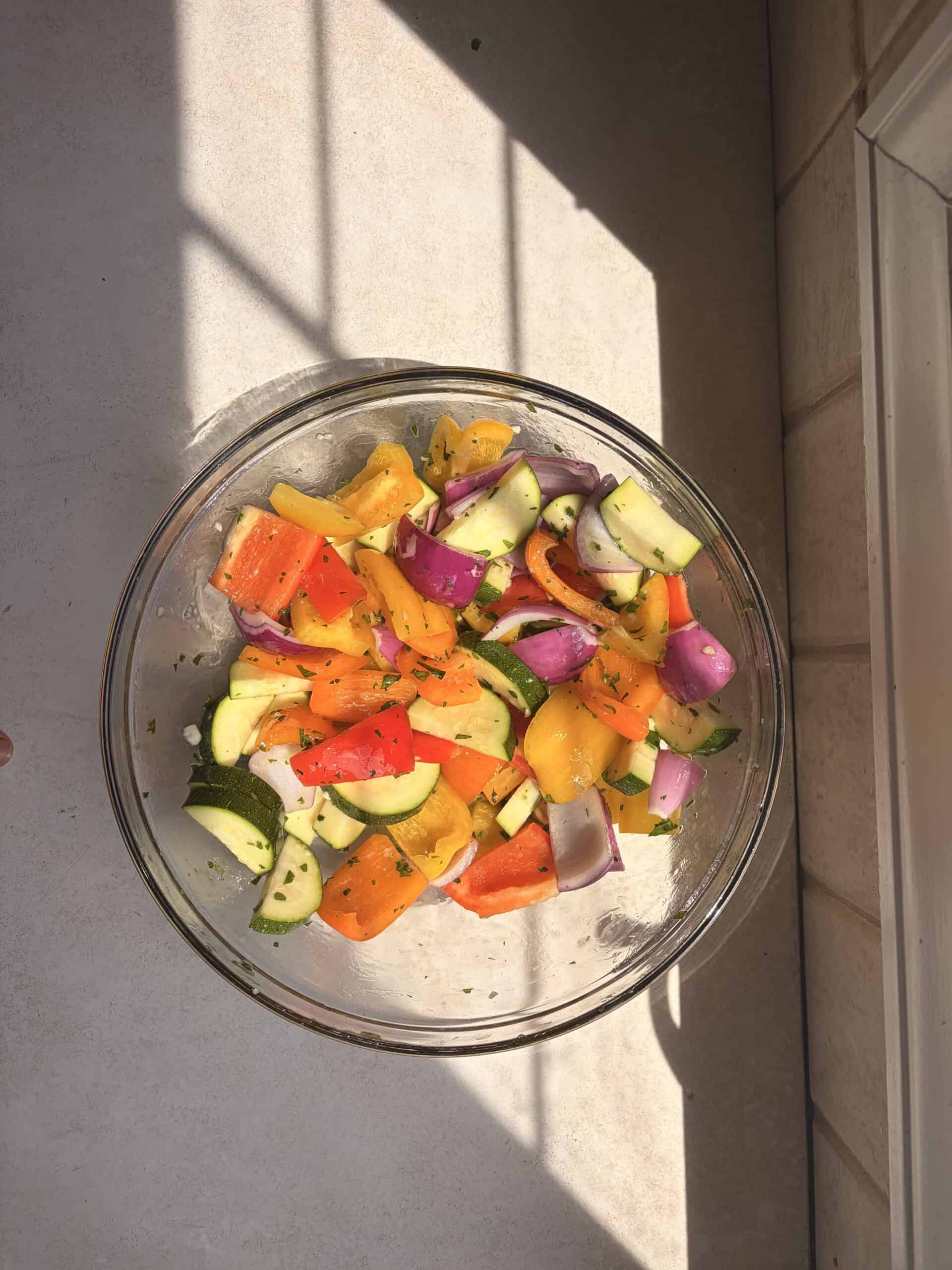 A glass bowl filled with chopped red, yellow, and orange bell peppers, red onions, and zucchini sits on a sunlit countertop near a window—perfect ingredients for assembling vibrant halloumi kebabs.