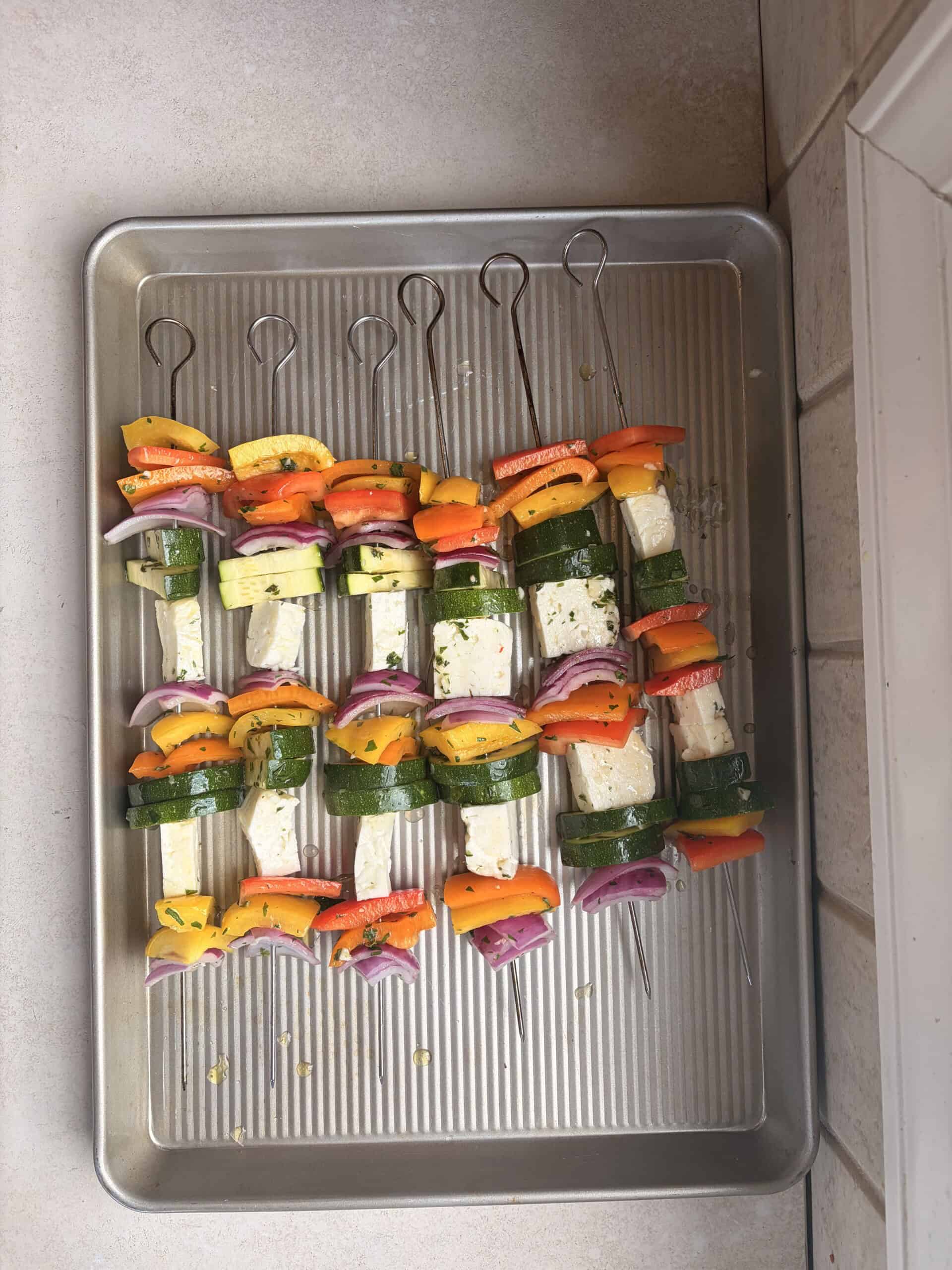 Four metal skewers with colorful chopped vegetables, including zucchini, red onion, yellow and orange bell peppers, and cubes of halloumi cheese, are arranged on a baking tray.