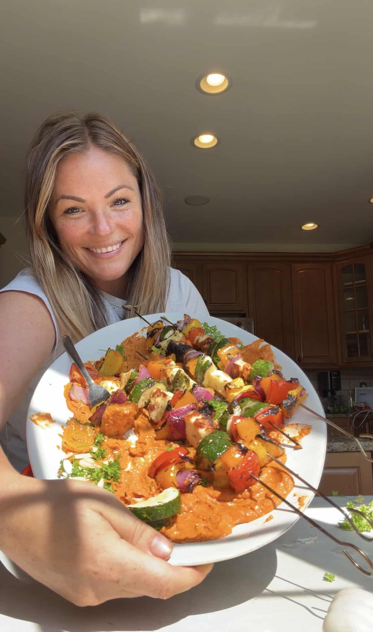 A smiling woman holds a plate of colorful halloumi kebabs and grilled vegetable skewers in a kitchen with wooden cabinets, bright lighting, and sunlight streaming in.