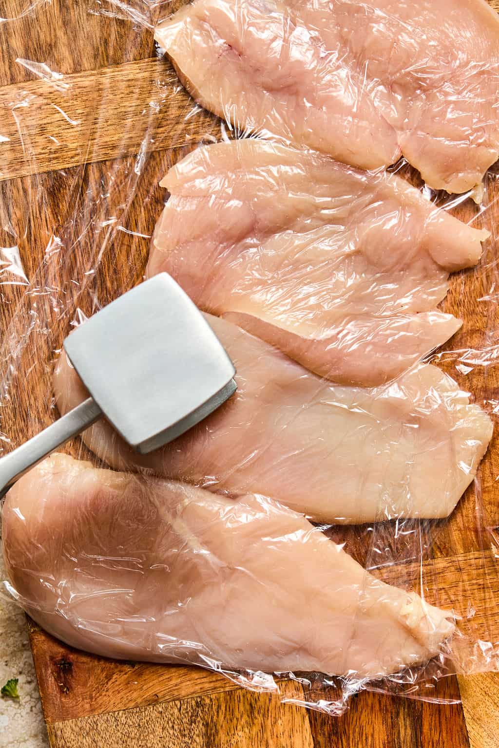 Raw chicken breasts on a wooden cutting board, covered with plastic wrap, being flattened with a metal meat mallet.