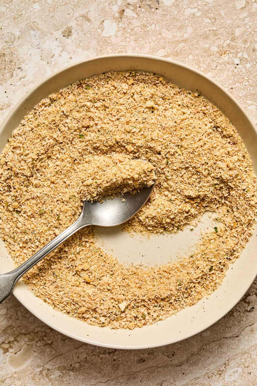 A metal spoon rests in a shallow bowl filled with seasoned breadcrumbs on a beige, textured surface.