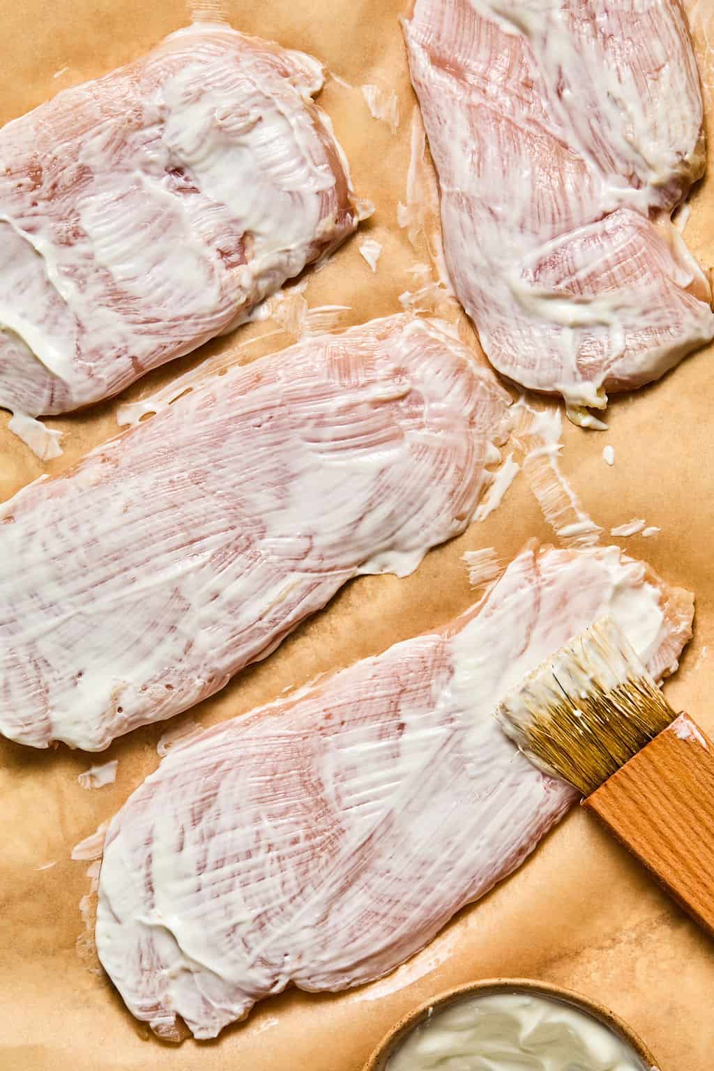 Raw chicken cutlets coated with a layer of Greek yogurt, spread using a wooden brush on brown parchment paper, with a small bowl of extra sauce nearby.