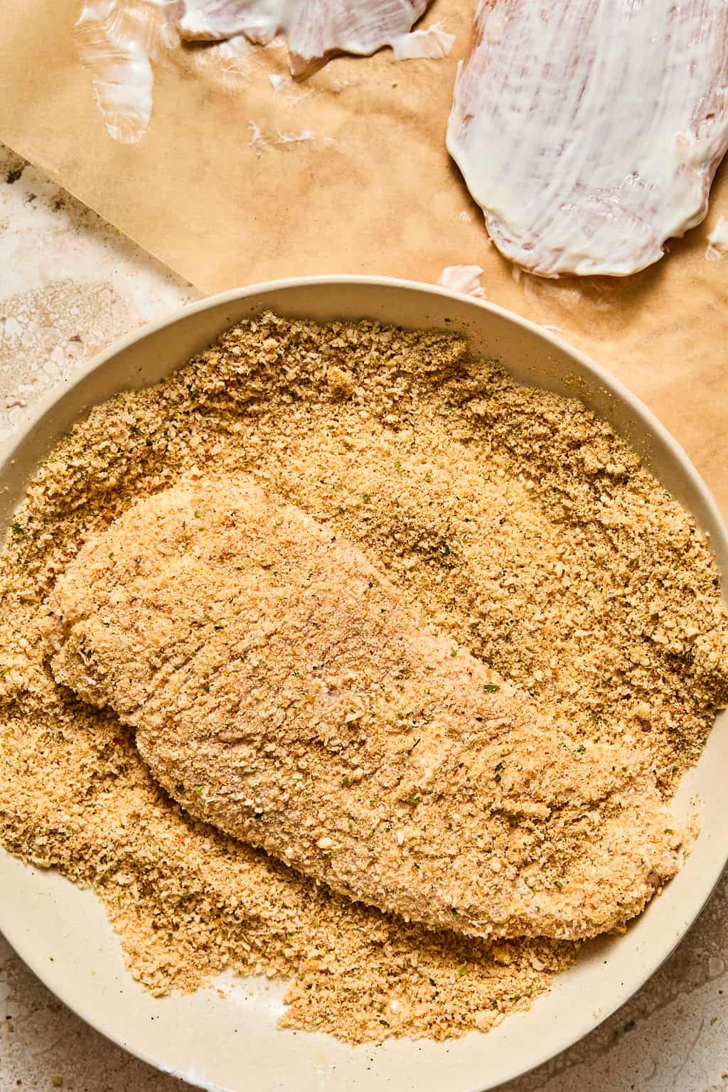 A raw, coated fillet is being breaded in seasoned crumbs on a plate. In the background, another fillet is covered in greek yogurt on brown parchment paper.