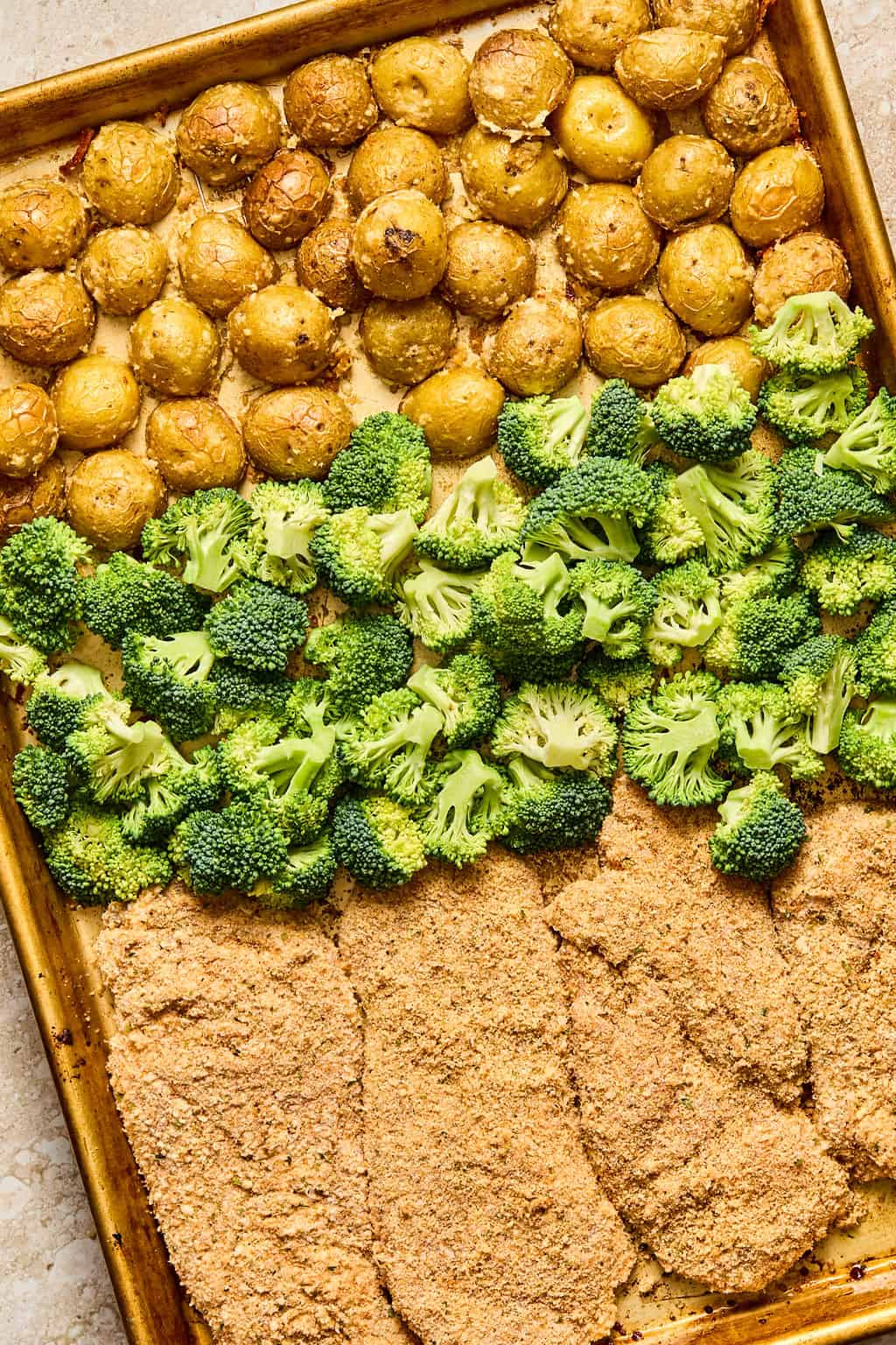 A sheet pan with rows of roasted baby potatoes, fresh broccoli florets, and breaded chicken cutlets, all arranged neatly and baked together.