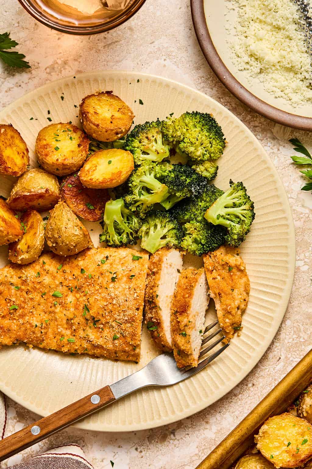 A plate with breaded chicken breast, roasted potatoes, and steamed broccoli. A fork rests on the plate with a slice of chicken. Parmesan cheese and parsley garnish are visible nearby.