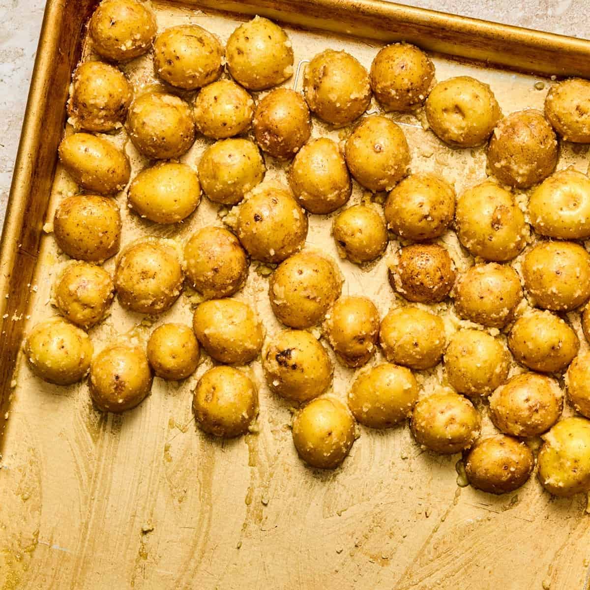 Golden roasted baby potatoes are spread out on a baking sheet. The potatoes are evenly coated with oil, parmesan, and salt, before baking.