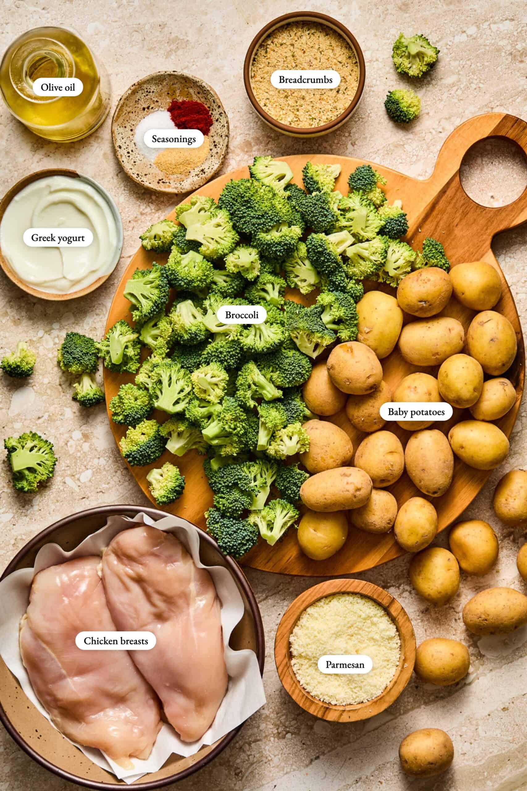 Ingredients on a counter: raw chicken breasts, baby potatoes, chopped broccoli, breadcrumbs, parmesan, Greek yogurt, olive oil, and seasonings, all labeled and displayed in bowls or on a wooden board.