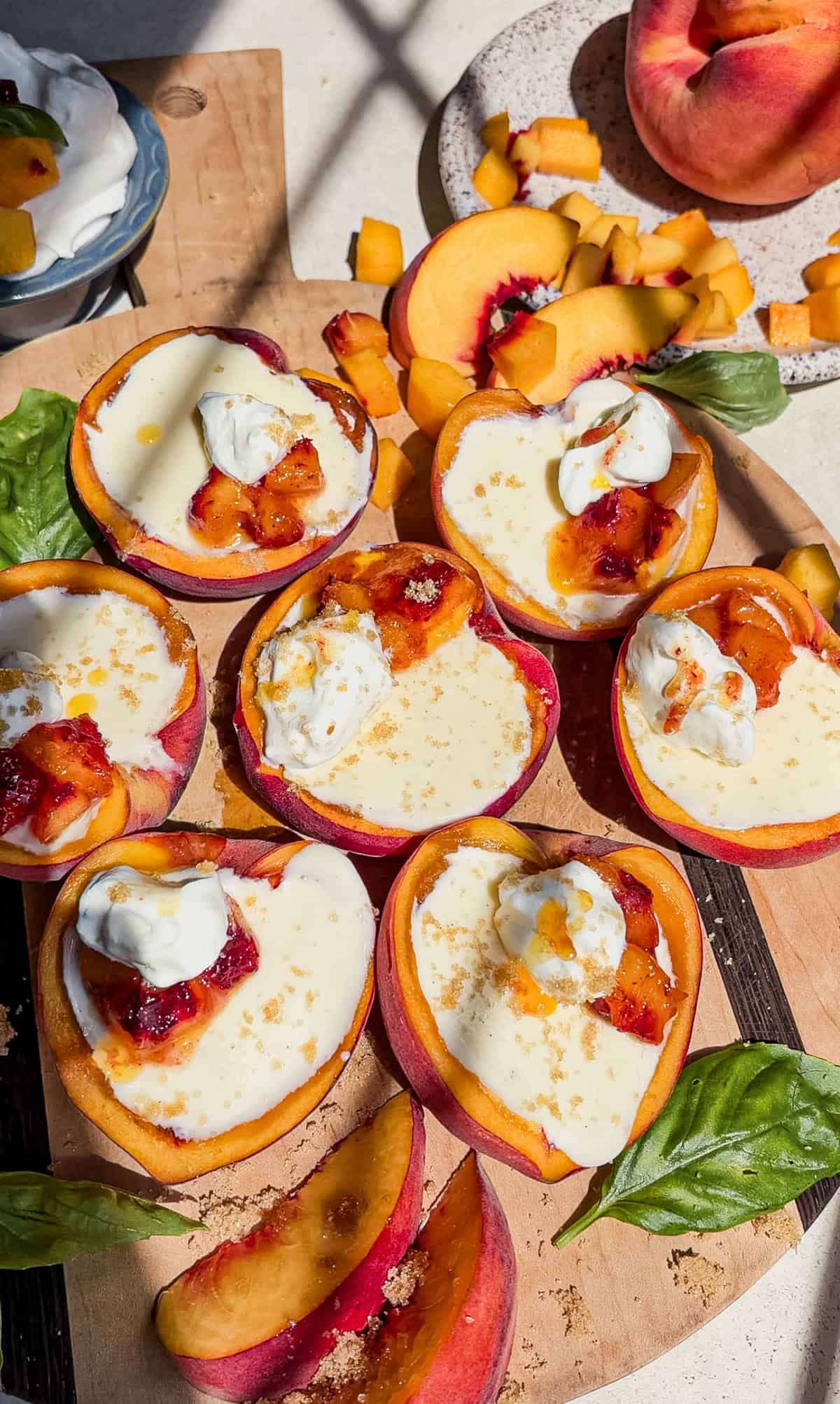 Halved peaches filled with posset cream and topped with peaches, and whipped cream are arranged on a cutting board with peach slices, diced peaches, basil leaves, and a bowl of whipped cream nearby. Sunlight creates bright highlights.