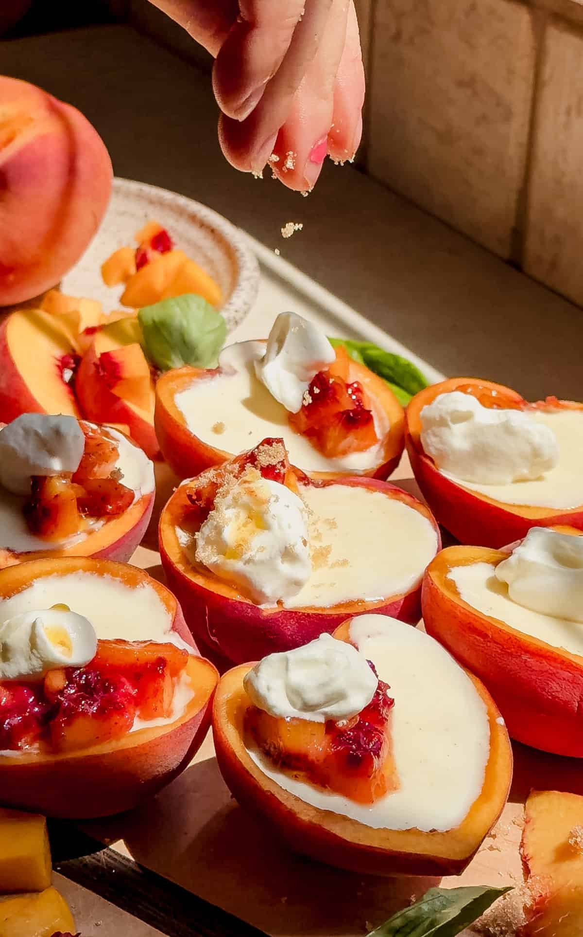 A hand sprinkles brown sugar over peach possets and topped with dollops of whipped cream and fruit preserves, arranged on a wooden surface in bright sunlight.