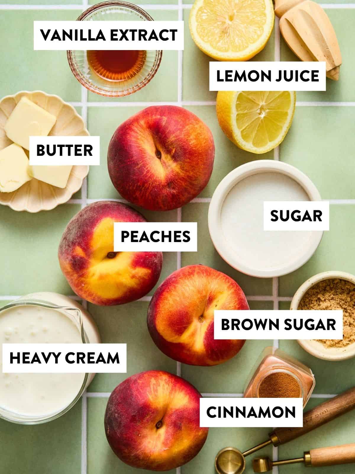 Top-down view of labeled ingredients on a green tiled surface for making peach posset: vanilla extract, lemon juice, butter, sugar, brown sugar, peaches, heavy cream, and cinnamon. Each ingredient is arranged neatly with clear labels.