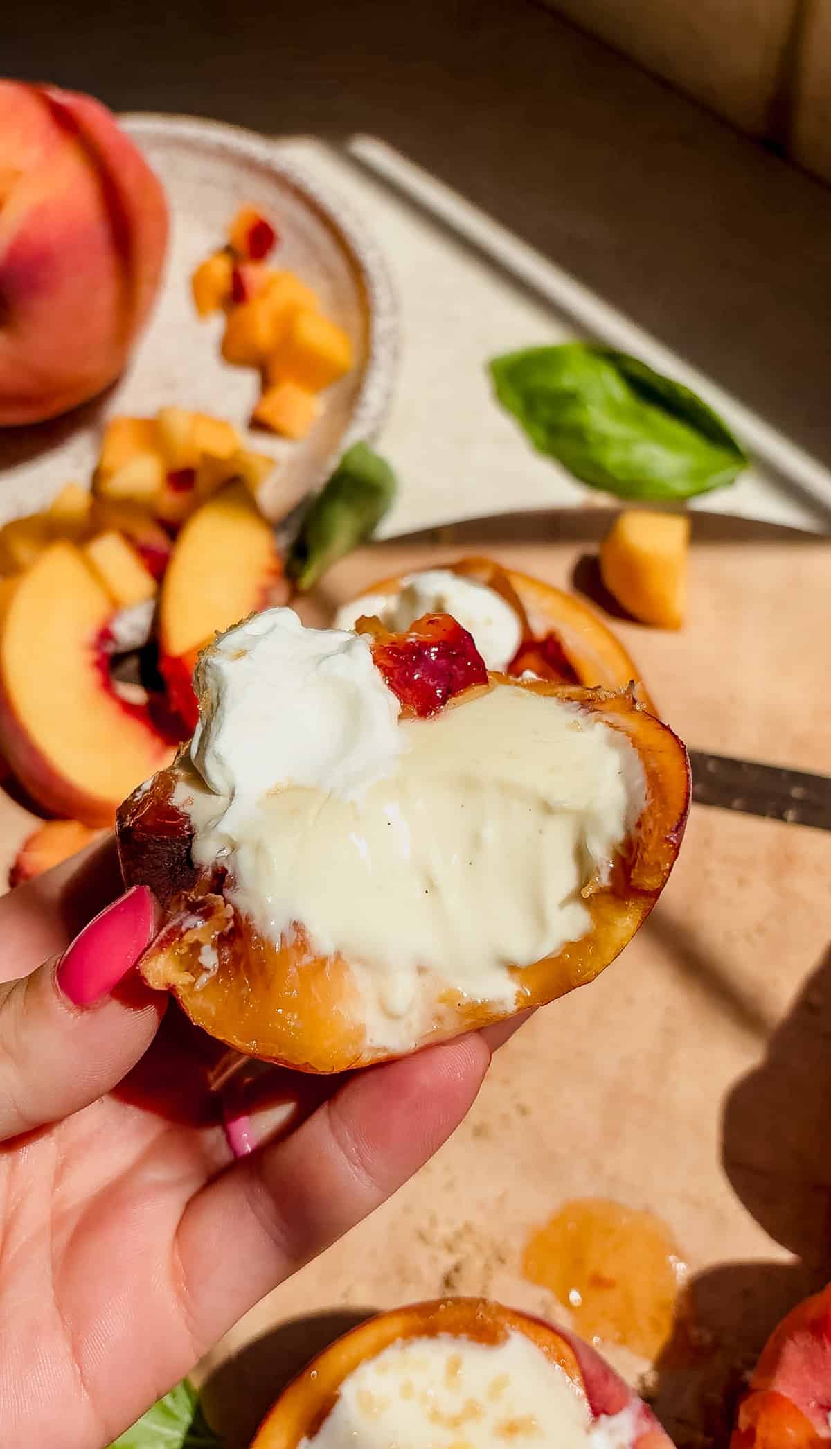 A hand with pink nails holds a peach half filled with posset. Sliced peaches, whipped cream, and basil leaves rest on a wooden cutting board in the bright sunlight.