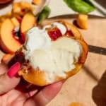 A hand holds a halved peach topped with whipped cream and creamy white sauce, with peach slices and a knife on a wooden cutting board in the background.