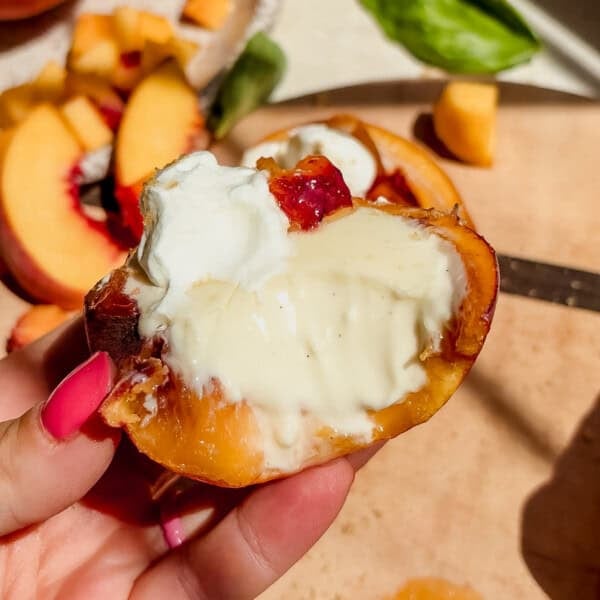 A hand holds a halved peach topped with whipped cream and creamy white sauce, with peach slices and a knife on a wooden cutting board in the background.