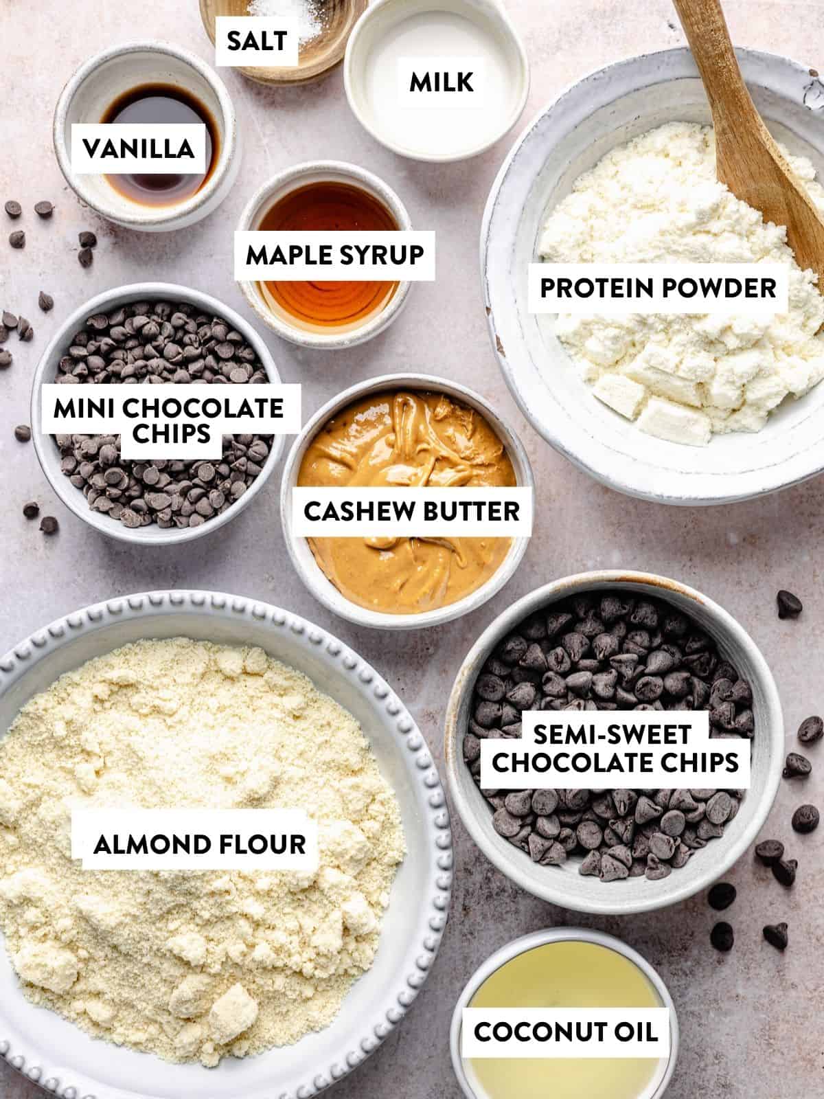 Bowls with labeled ingredients—salt, milk, vanilla, maple syrup, protein powder, mini chocolate chips, cashew butter, semi-sweet chocolate chips, almond flour, and coconut oil—are all arranged on a light surface for making delicious protein cookie dough bark.