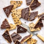 Triangular pieces of chocolate-topped protein cookie dough bark with sea salt are scattered on parchment paper. A knife with a wooden handle is placed among the pieces.