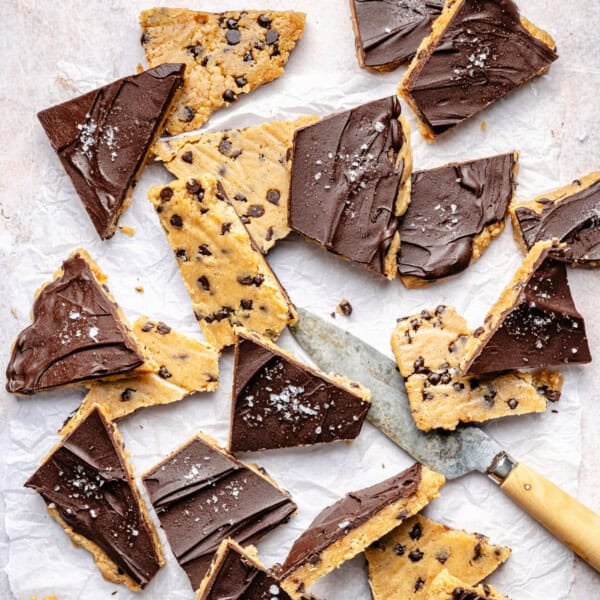 Triangular pieces of chocolate-topped protein cookie dough bark with sea salt are scattered on parchment paper. A knife with a wooden handle is placed among the pieces.